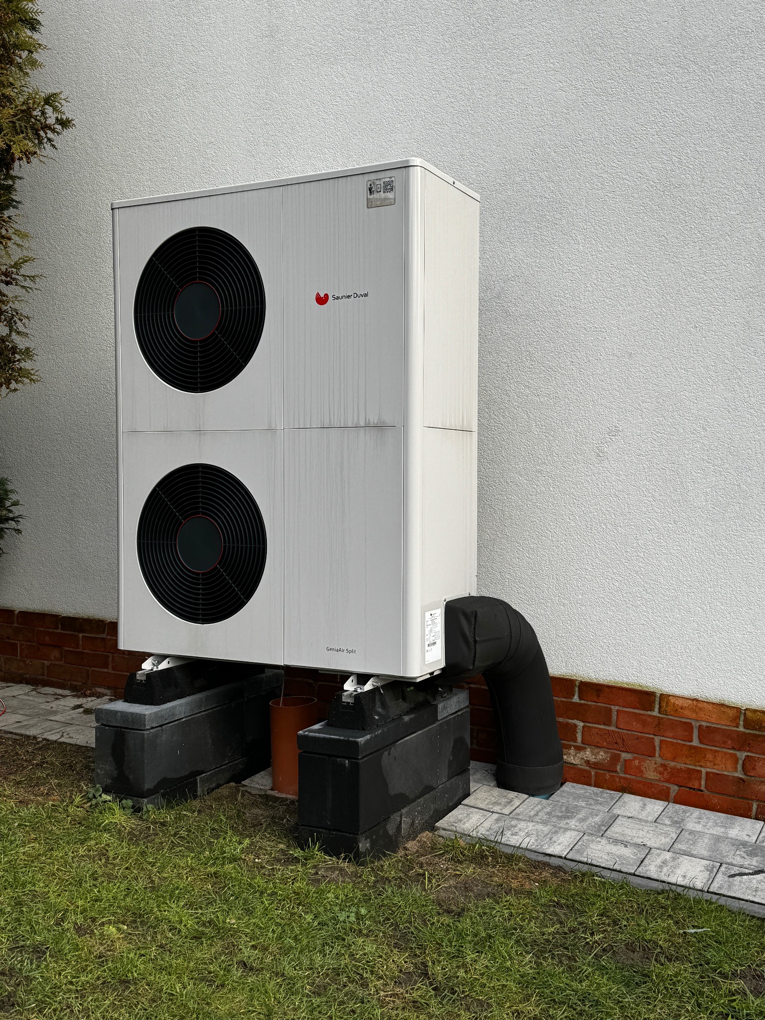 Biała pompa ciepła Saunier Duval GeniaAir Split, zamontowana na betonowych blokach przy ścianie budynku z ceglaną podmurówką i trawnikiem.