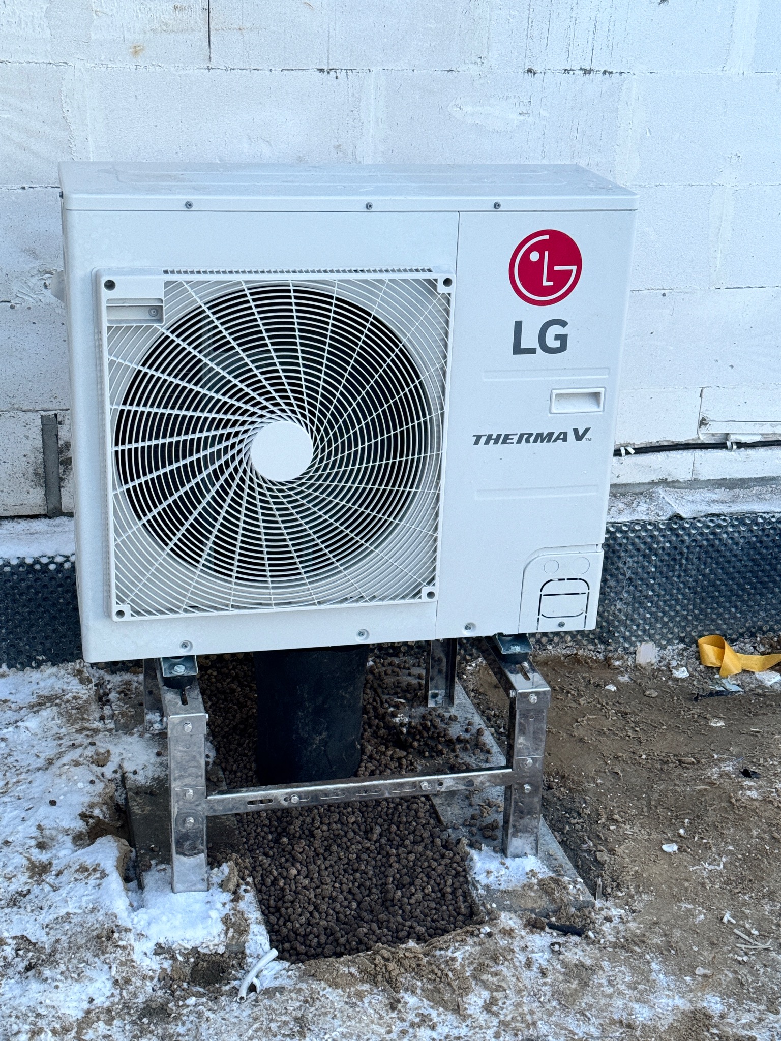 Biała pompa ciepła LG Therma V zamontowana na metalowym stelażu przy ścianie z betonu komórkowego. Wokół widoczny śnieg i brązowe granulki izolacyjne.
