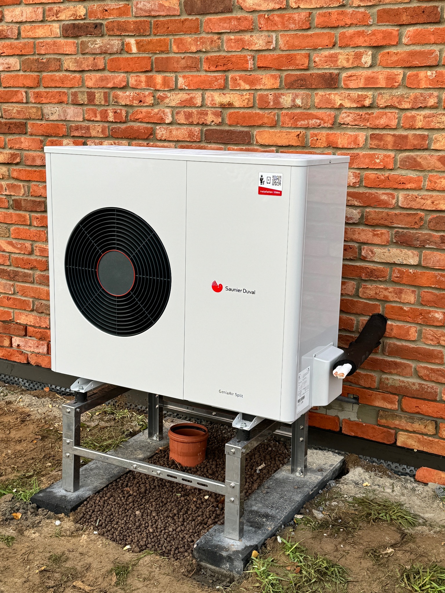 Biała pompa ciepła Saunier Duval GeniaAir Split, zamontowana na metalowej konstrukcji przy ceglanej ścianie w Gdańsku. Widoczne rury i elementy instalacji.