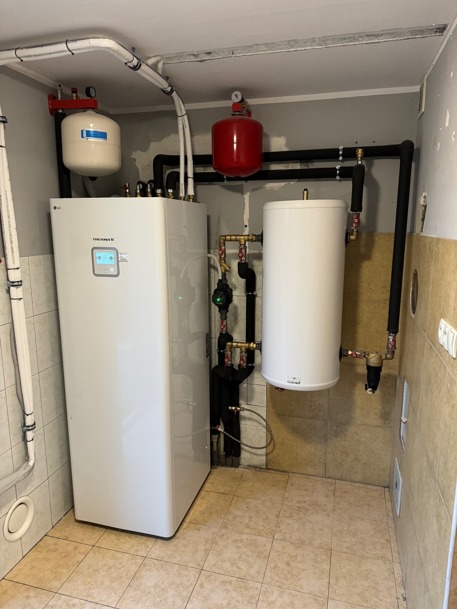 Zmontowana instalacja z pompą ciepła LG Therma V, zbiornikiem Galmet i rurami w piwnicy. Widoczne zawory, manometry i elementy mocujące. Beżowe płytki na ścianach i podłodze.