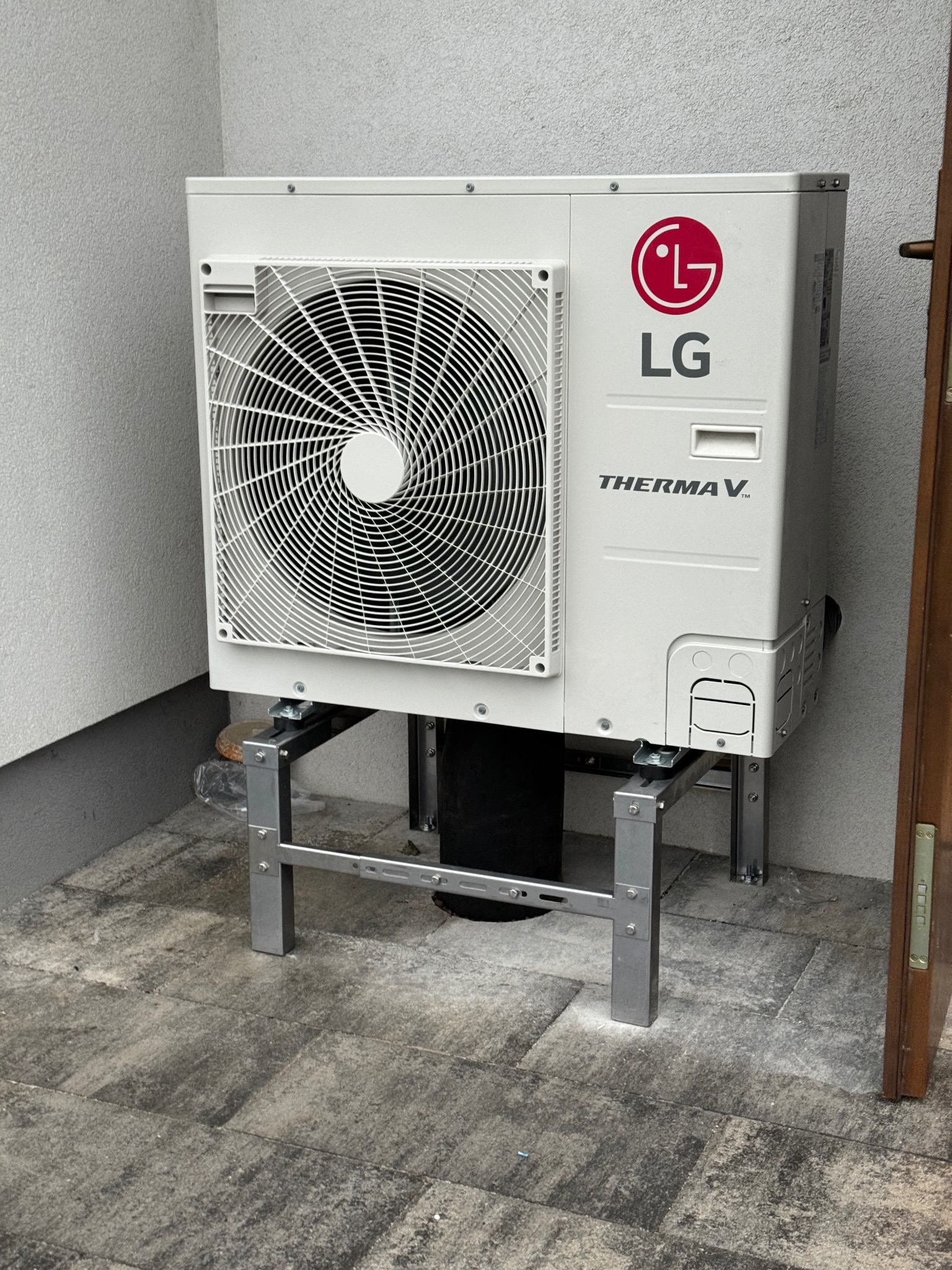 Biała pompa ciepła LG Therma V zamontowana na metalowym stelażu, ustawiona na szarym bruku przy ścianie budynku. Widoczny fragment brązowych drzwi.