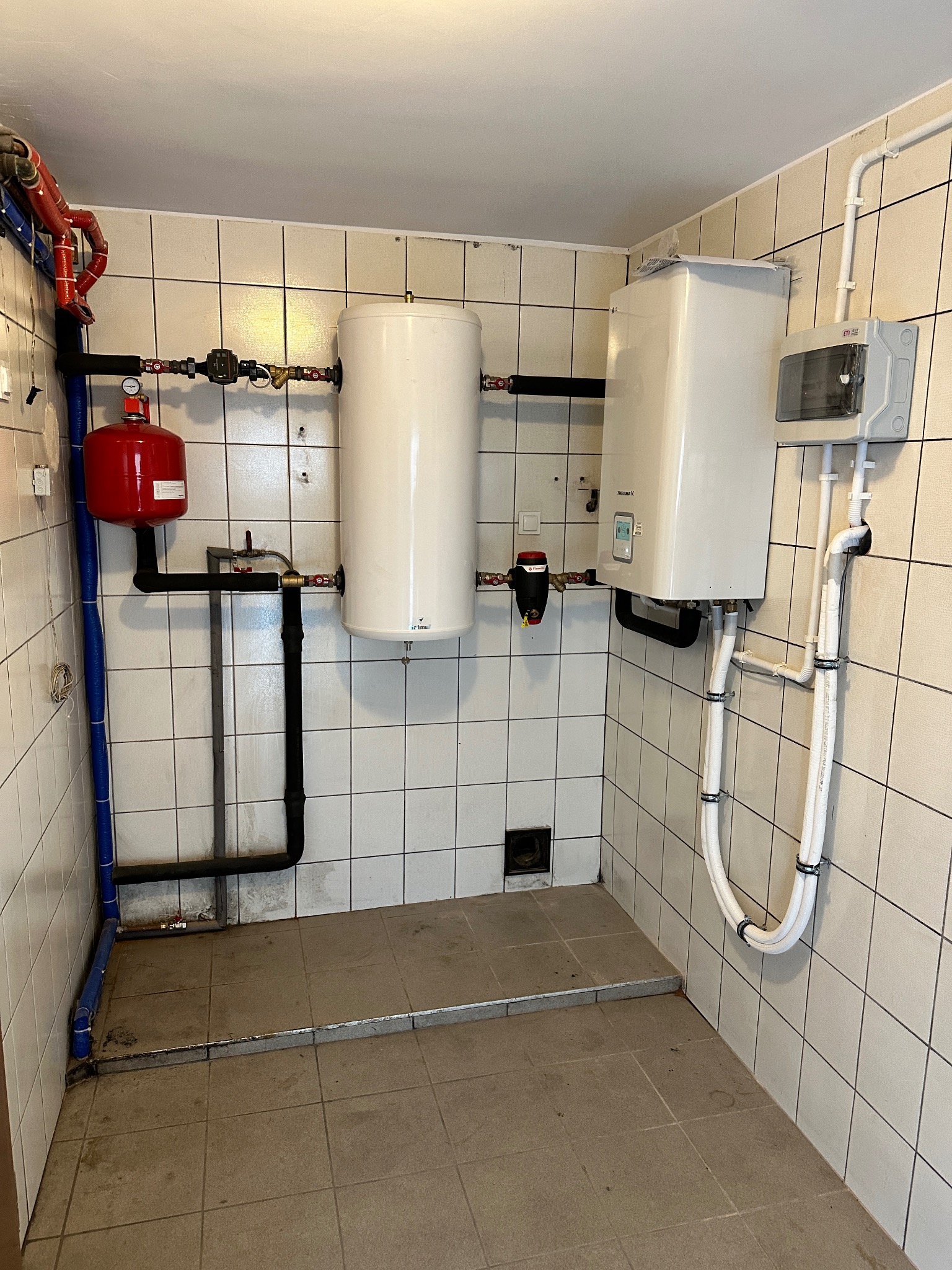 Biały bojler i piec gazowy Thermava V zamontowane na ścianie z białych płytek, z widocznymi rurami i zaworami. Instalacja w pomieszczeniu z podłogą wyłożoną płytkami.