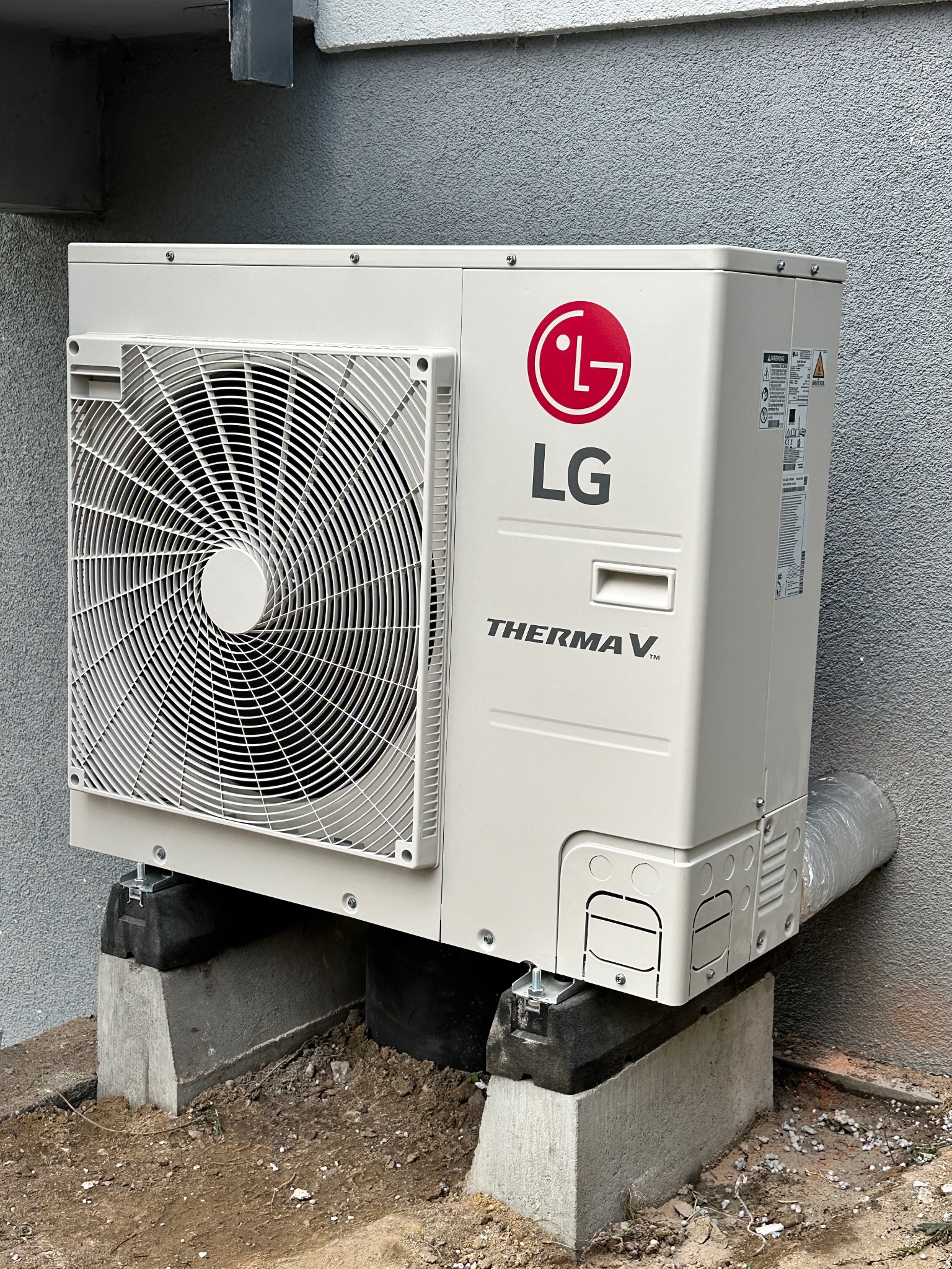 Biała pompa ciepła LG Therma V, zamontowana na betonowych podstawach przy szarej ścianie budynku. Widoczny wentylator i rura odprowadzająca.