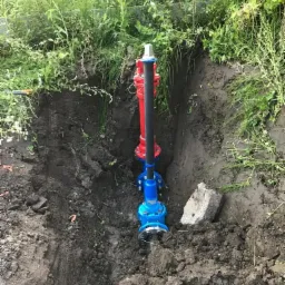 Nowe przyłącze wodociągowe w wykopie, z widocznymi elementami złączne w kolorze niebieskim i czerwonym, otoczone ziemią i fragmentami betonu.