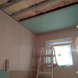 USLUGI REMONTOWO BUDOWLANE - Montaż sufitu podwieszanego z płyty gipsowej: widoczne profile, częściowo zamontowana płyta o zielonym odcieniu, drabina i okno w tle.