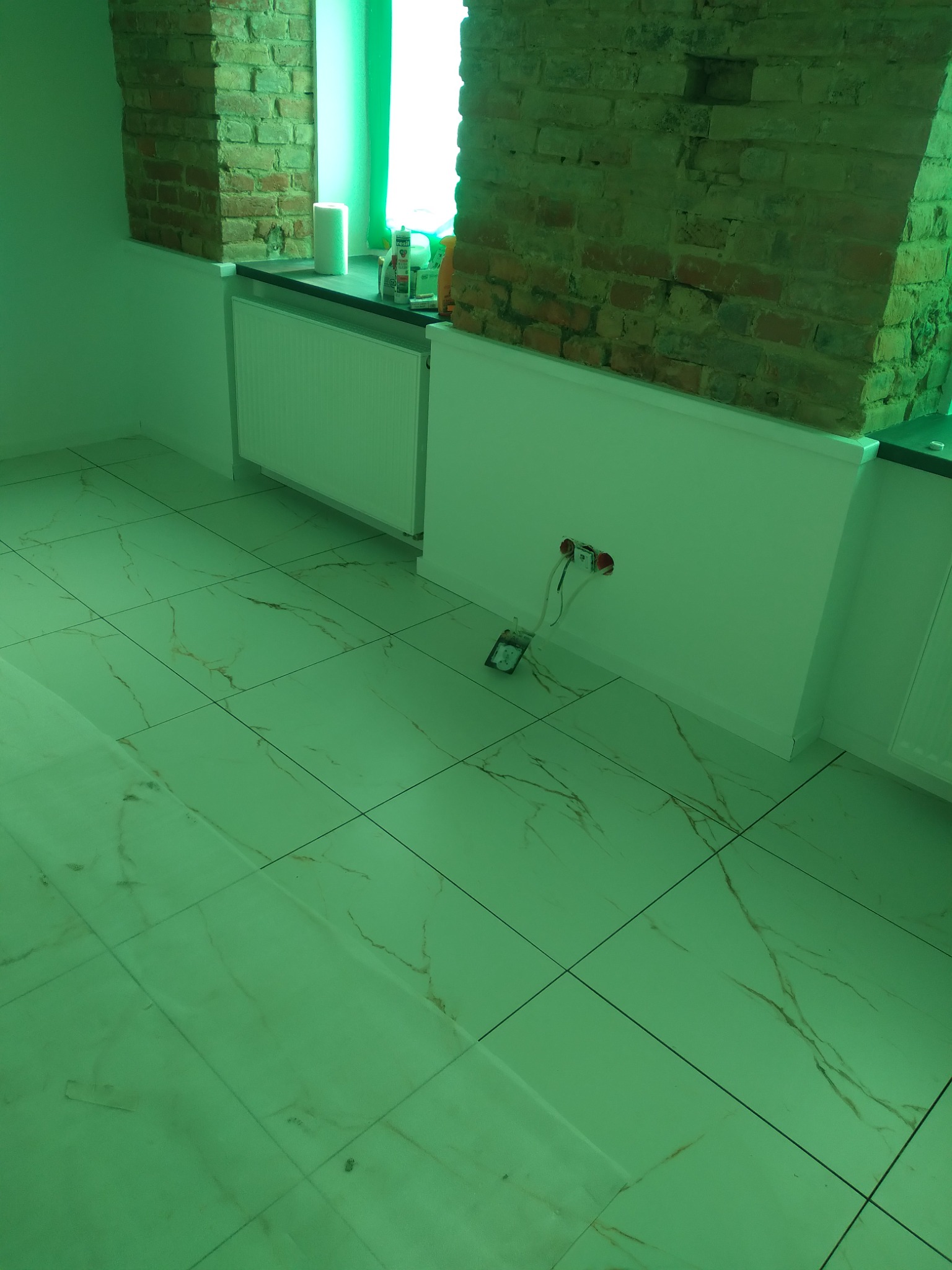 Wnętrze w trakcie prac wykończeniowych: widoczna nowa marmurowa podłoga, fragment ściany z odkrytą cegłą, grzejnik oraz puszka elektryczna z wystającymi przewodami.