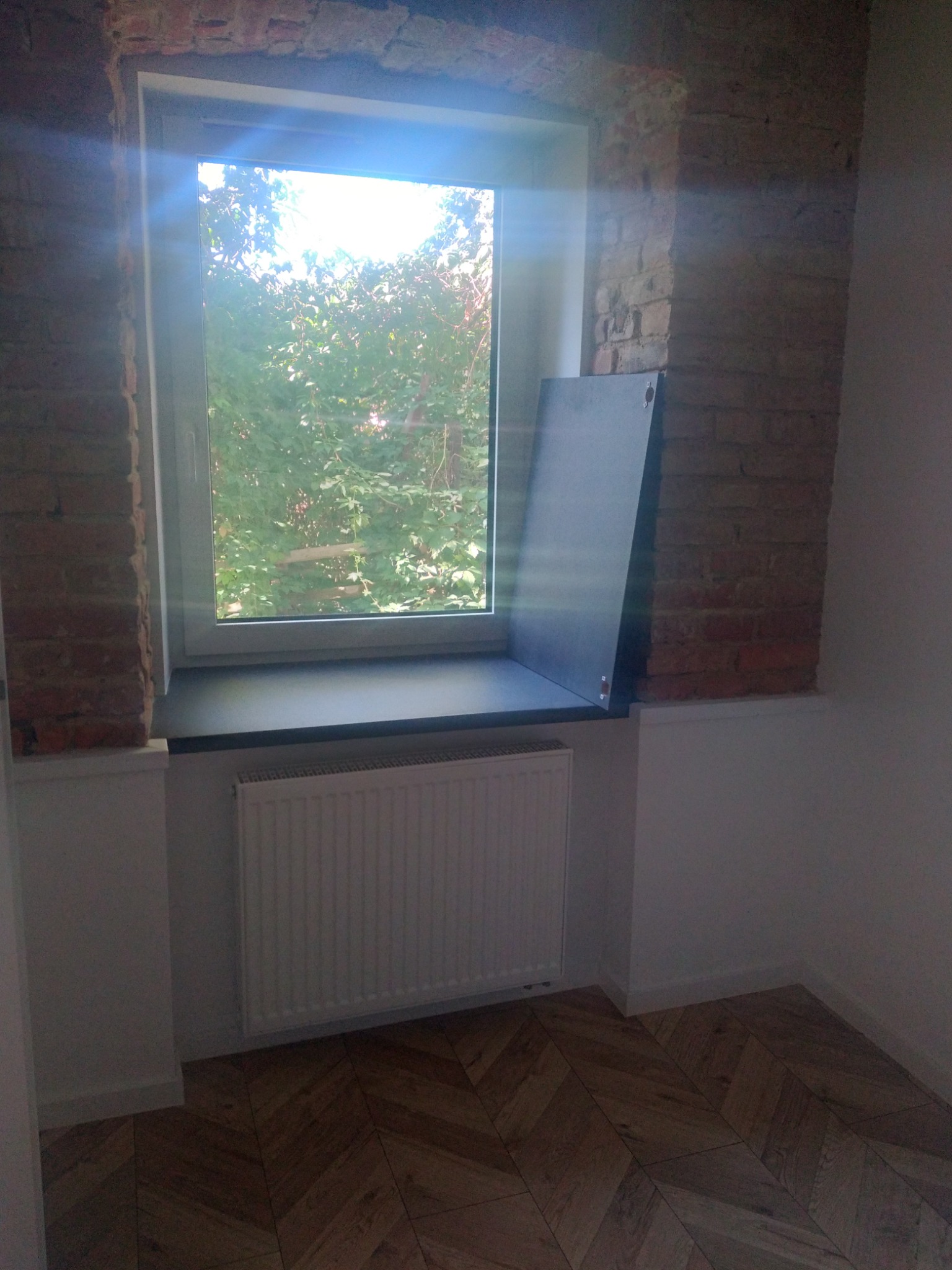 Pokój po remoncie z oknem, odsłoniętą cegłą, nowym parapetem i grzejnikiem, oraz parkietem ułożonym we wzór jodełki.