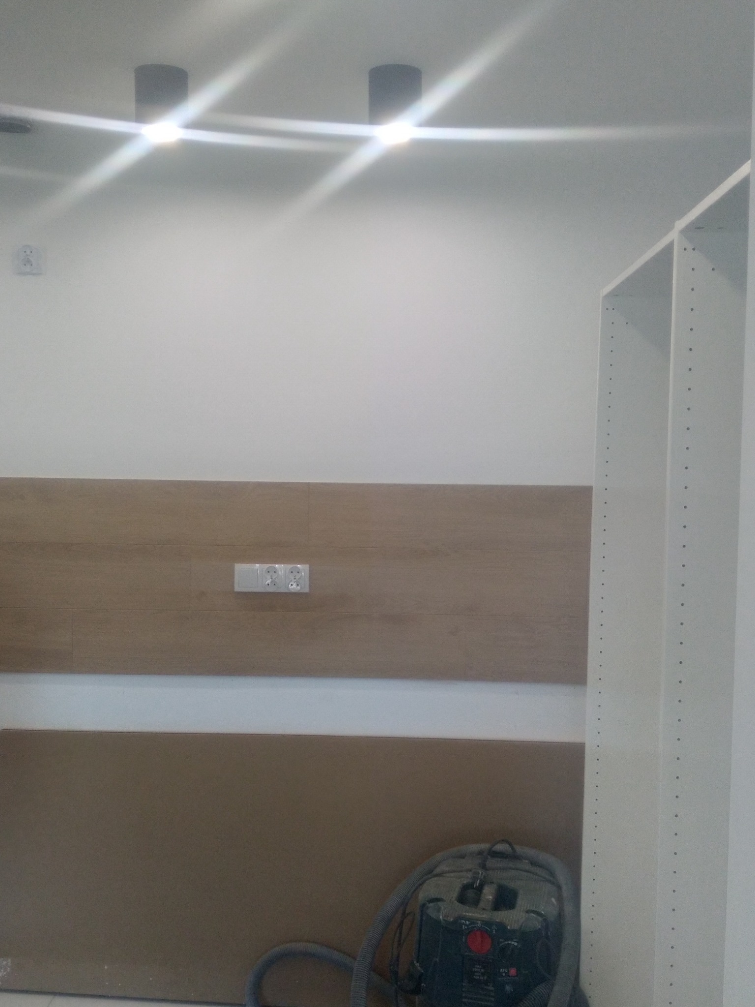 Fragment wnętrza w trakcie prac wykończeniowych: widoczna ściana z zamontowanym panelem drewnopodobnym, gniazdka elektryczne, fragment regału z otworami oraz odkurzacz przemysłowy w dolnym rogu.