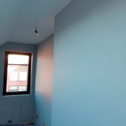 Świeżo pomalowane na szaro-niebieski kolor pomieszczenie ze skośnym sufitem, widoczne przygotowania do montażu armatury przy oknie oraz niezabezpieczone kable elektryczne.