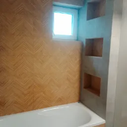 Biała wanna obudowana płytkami imitującymi drewno ułożonymi we wzór jodełki, z małym oknem nad wanną i wnękami w szarej ścianie z płytek.