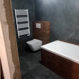 Nowoczesna aranżacja łazienki w trakcie wykańczania: grafitowe płytki, wanna obudowana drewnopodobną okładziną, podwieszana toaleta i drabinka grzejnikowa na ścianie, widoczne niedokończone...