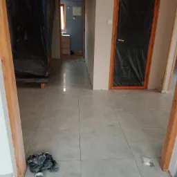 Szare płytki podłogowe ułożone w korytarzu, z widocznymi drzwiami zabezpieczonymi folią malarską, fragmentem ściany z gniazdkiem elektrycznym i paczkami z klejem do płytek.