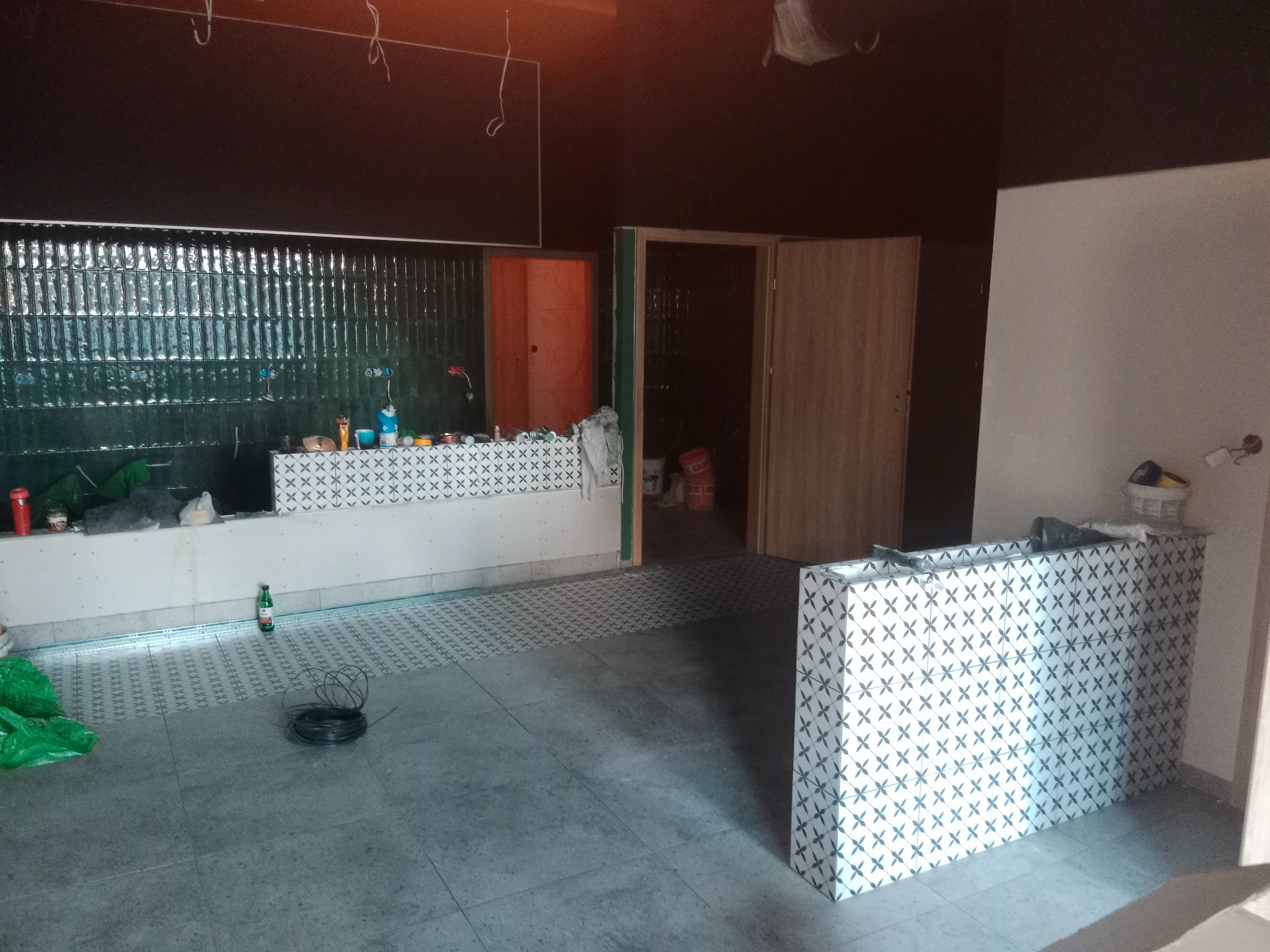 Wnętrze w trakcie prac wykończeniowych: ułożona szara podłoga z płytek, fragment ściany z zielonymi płytkami, przygotowane miejsce pod blat z akcesoriami, widoczne kable i butelka. Czarno...