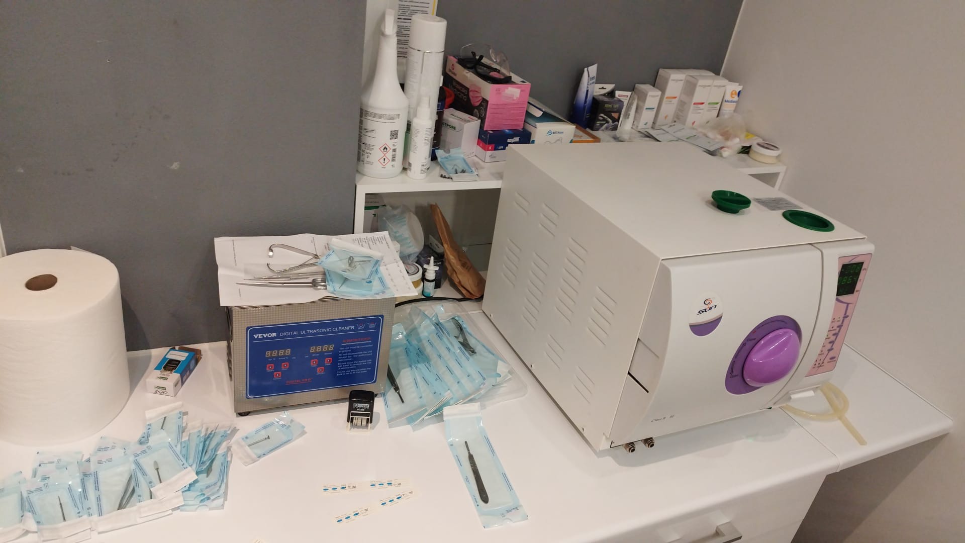 Sterylizator parowy, myjka ultradźwiękowa i pakowane narzędzia kosmetyczne na białym stole, przygotowane do zabiegów.