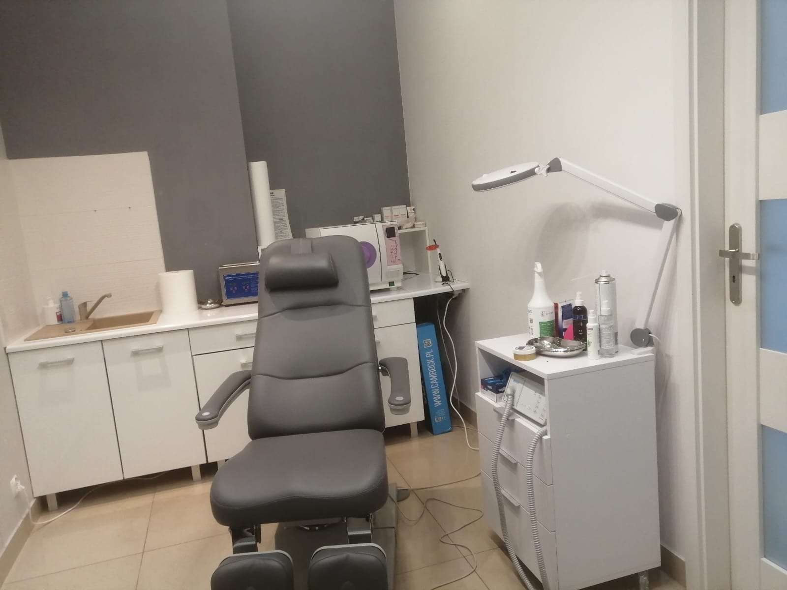 Gabinet podologiczny: Fotel do pedicure z regulowanym zagłówkiem, obok szafka z lampą powiększającą i preparatami kosmetycznymi, w tle umywalka i sterylizator.