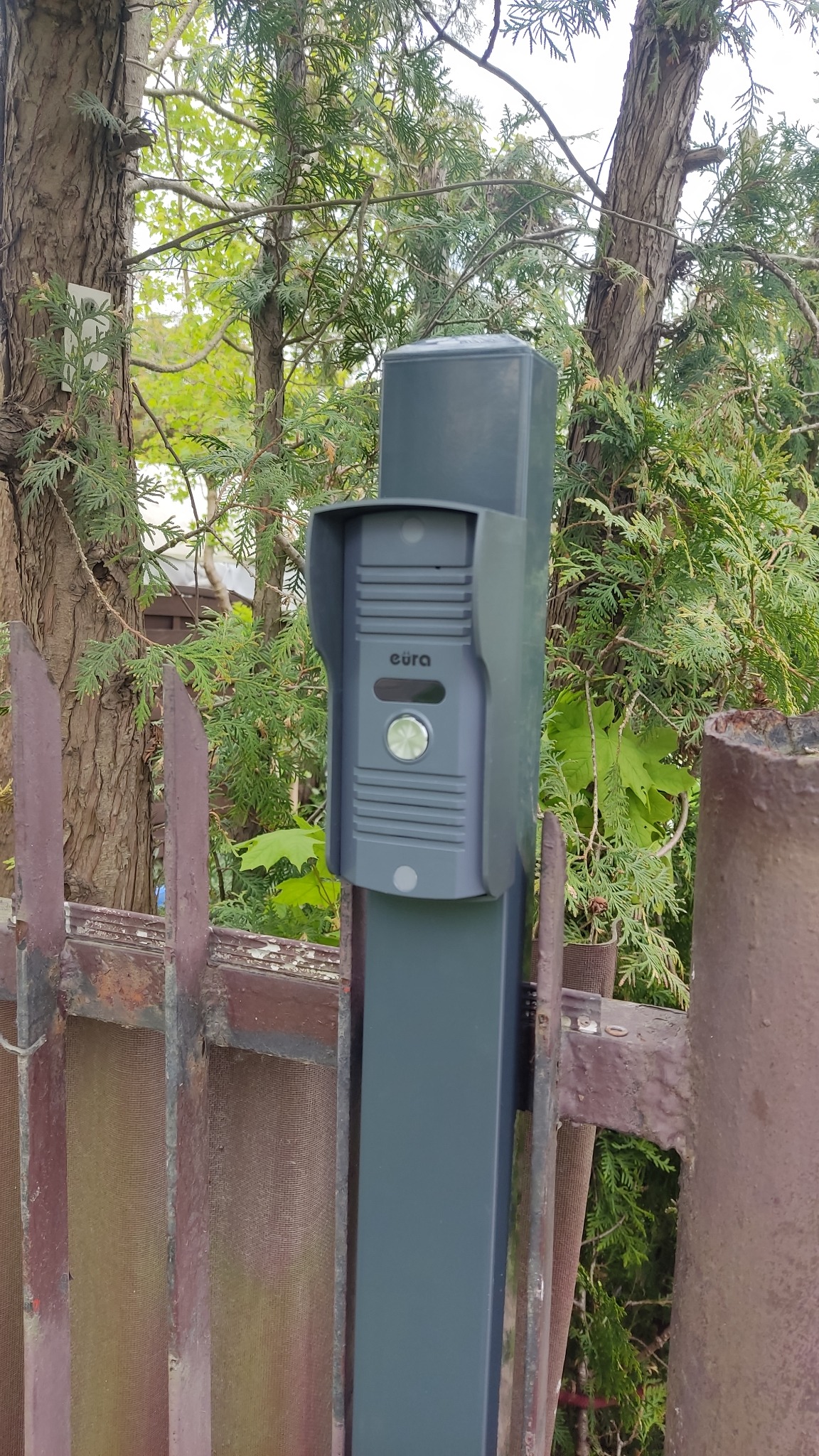 Szary domofon marki Eura zamontowany na metalowym słupku, widoczny na tle drzew i fragmentu zardzewiałego ogrodzenia.