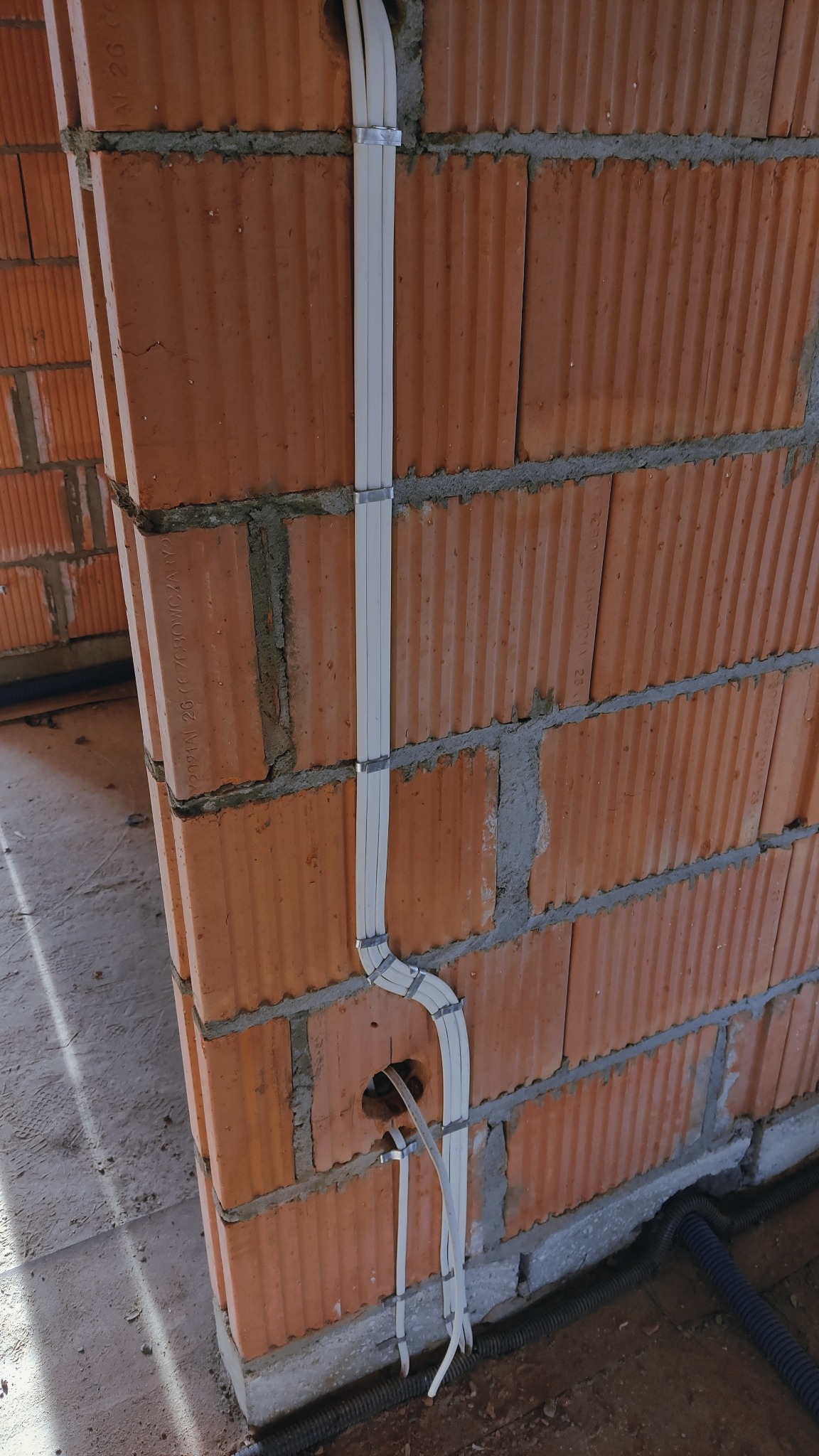 Białe przewody elektryczne prowadzone wtynkowo, przymocowane metalowymi uchwytami do ściany z czerwonej cegły dziurawki, z widocznym otworem w ścianie i czarną rurą karbowaną przy podłodze.