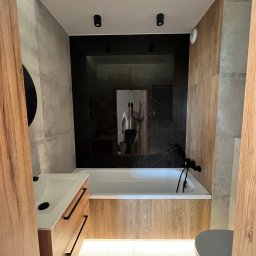 Art_HomeSERWIS Vitalii Kvasniuk - Nowoczesna łazienka z czarną armaturą, podświetleniem LED pod wanną i szafką umywalkową, oraz lustrzaną ścianą z odbiciem osoby.