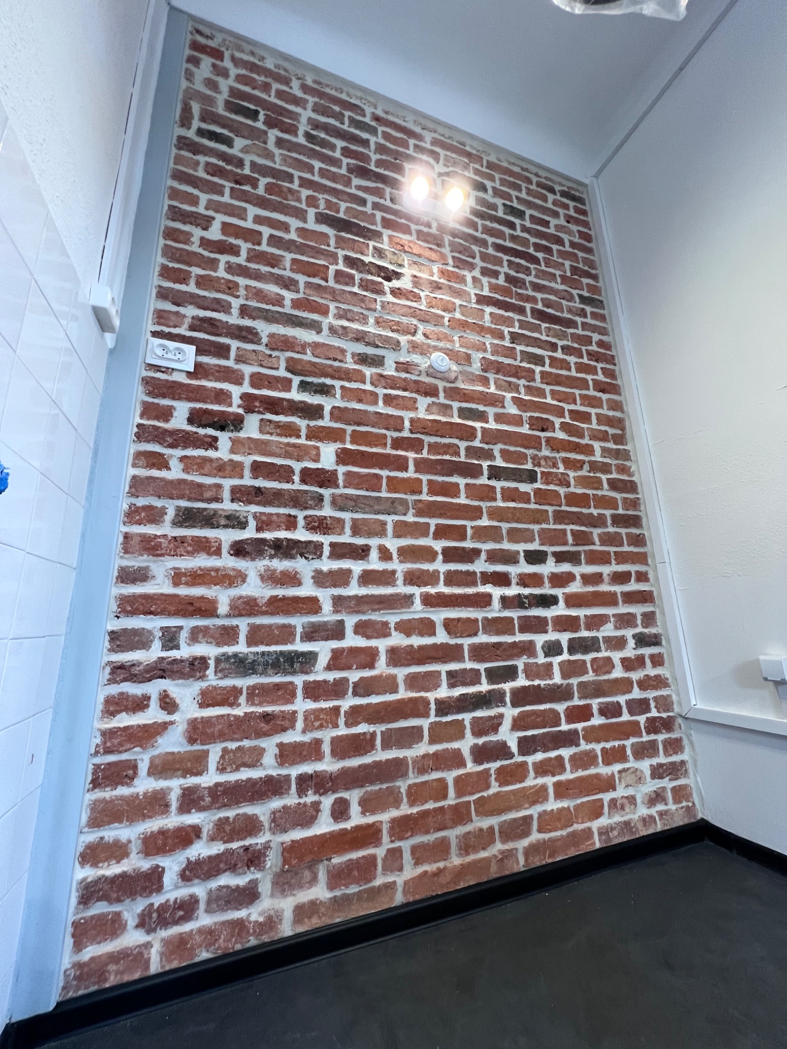 Ściana z czerwonej cegły z białymi fugami, oświetlona dwoma reflektorami, z widocznym gniazdkiem elektrycznym i fragmentem białej ściany z płytek ceramicznych po lewej stronie.