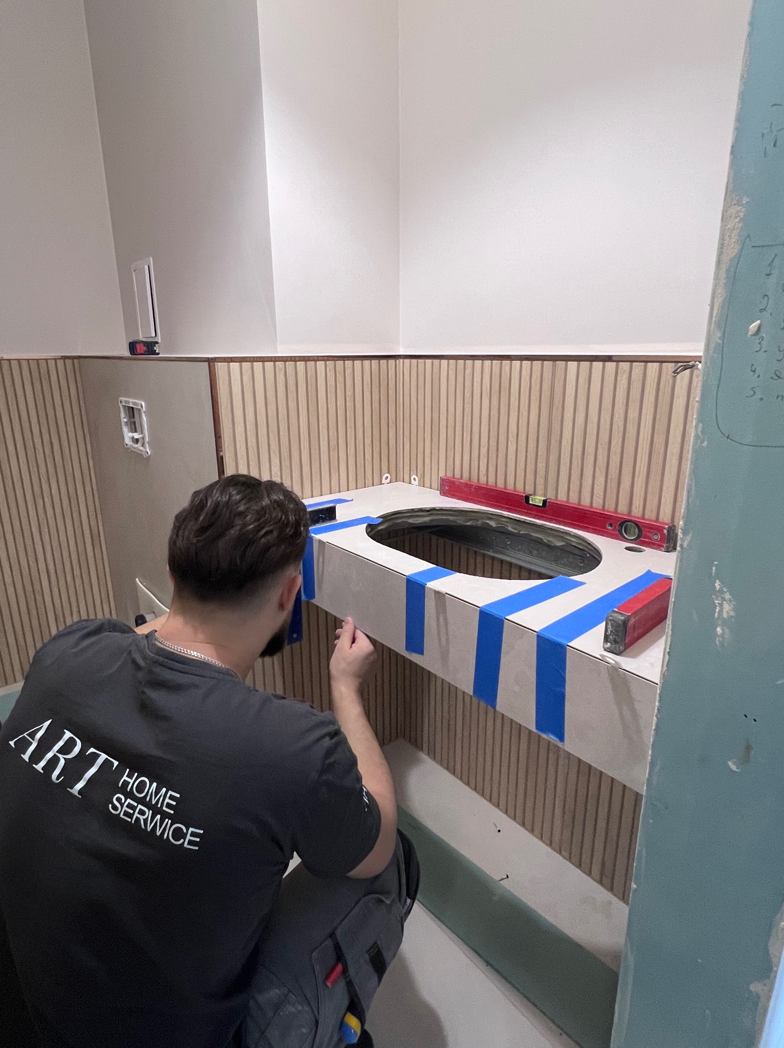Instalacja umywalki w łazience: pracownik w koszulce ART HOME SERVICE poziomuje blat z otworem pod umywalkę, zabezpieczony niebieską taśmą malarską, na tle drewnianej boazerii.