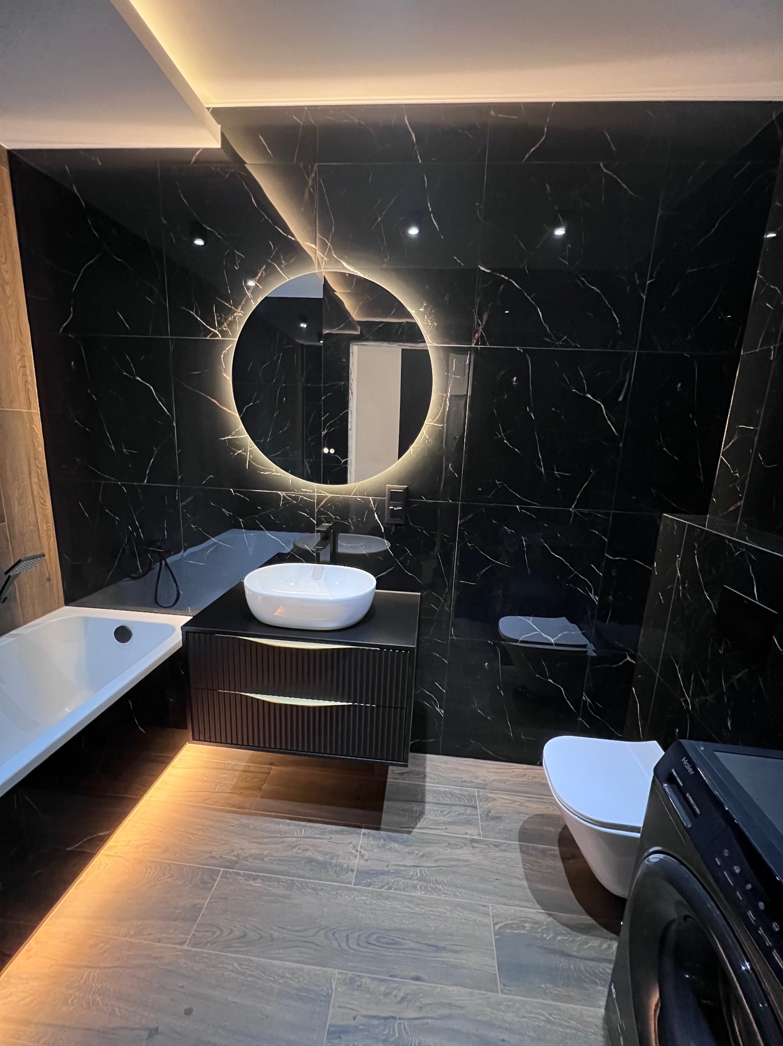 Elegancka łazienka z czarnymi płytkami imitującymi marmur, okrągłym lustrem podświetlanym LED, białą umywalką nablatową, drewnopodobną podłogą i widoczną pralką z przodu.