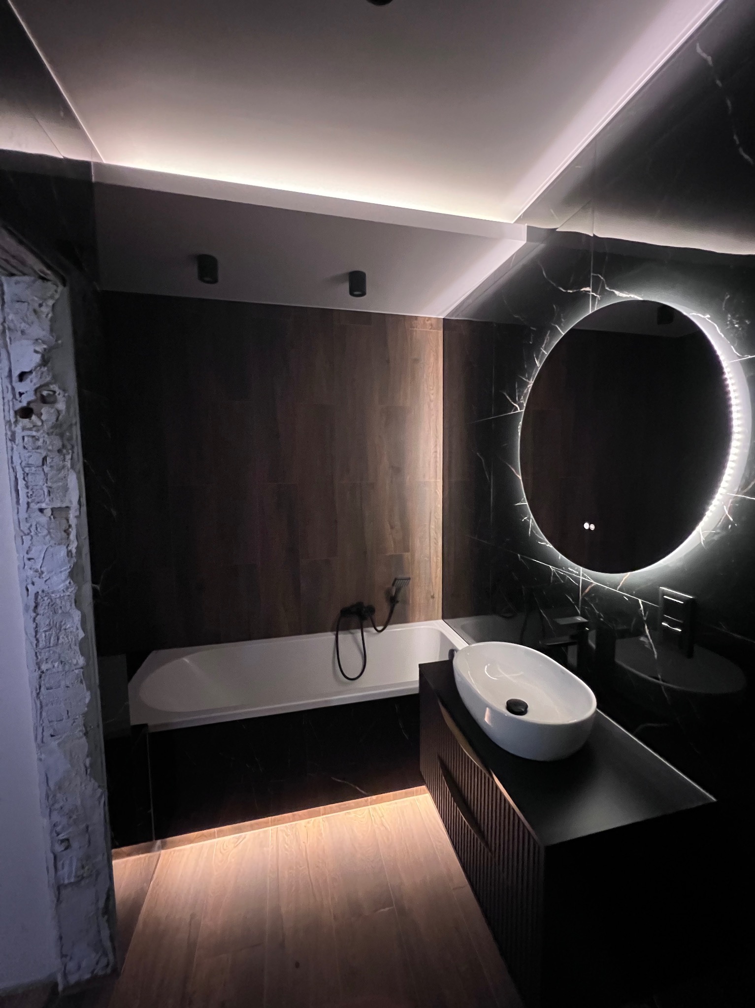 Nowoczesna łazienka z czarnymi marmurowymi płytkami, okrągłym lustrem z podświetleniem LED, białą owalną umywalką nablatową na czarnej szafce i podświetlaną wanną, widoczny fragment ściany...