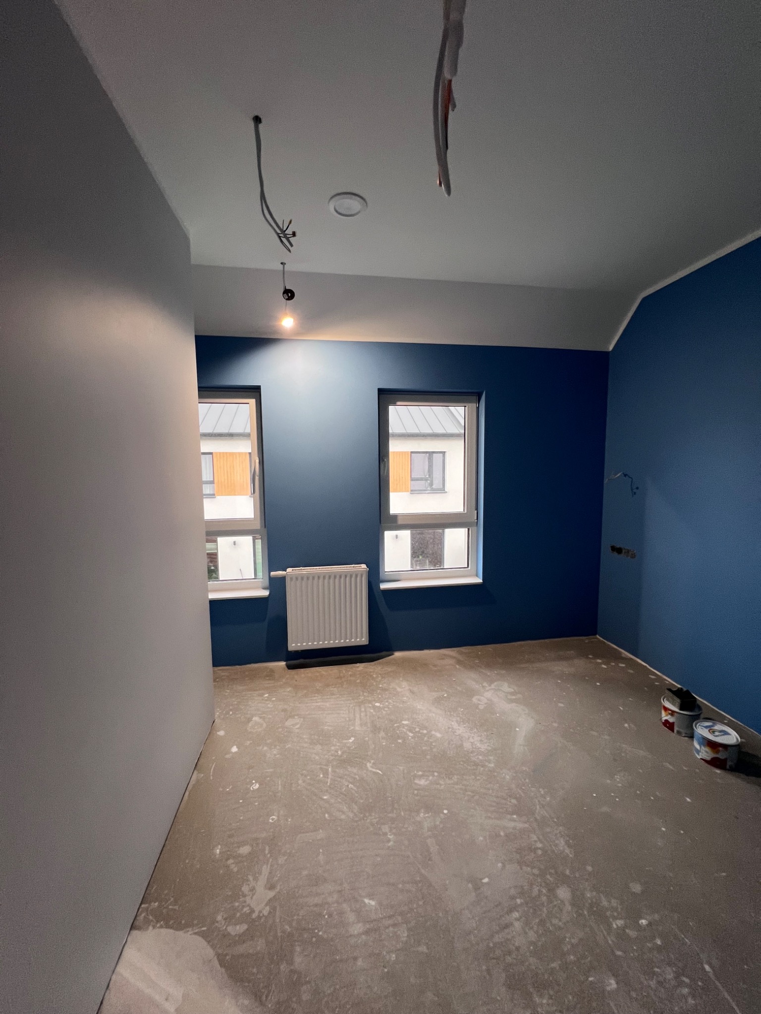 Pokój w trakcie malowania w Warszawie, ściany pomalowane na niebiesko, jedna ściana szara, widoczne dwa okna z grzejnikami, na podłodze ślady farby i puszki z farbą.