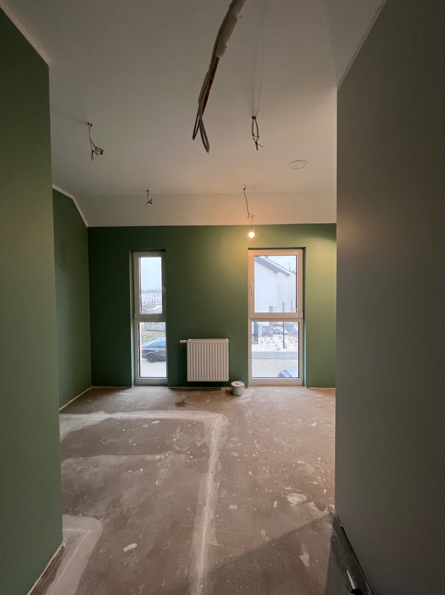 Pokój po malowaniu ścian na zielono, widoczne przygotowane podłoże pod wylewkę, wystające przewody elektryczne z sufitu, grzejnik i okna.