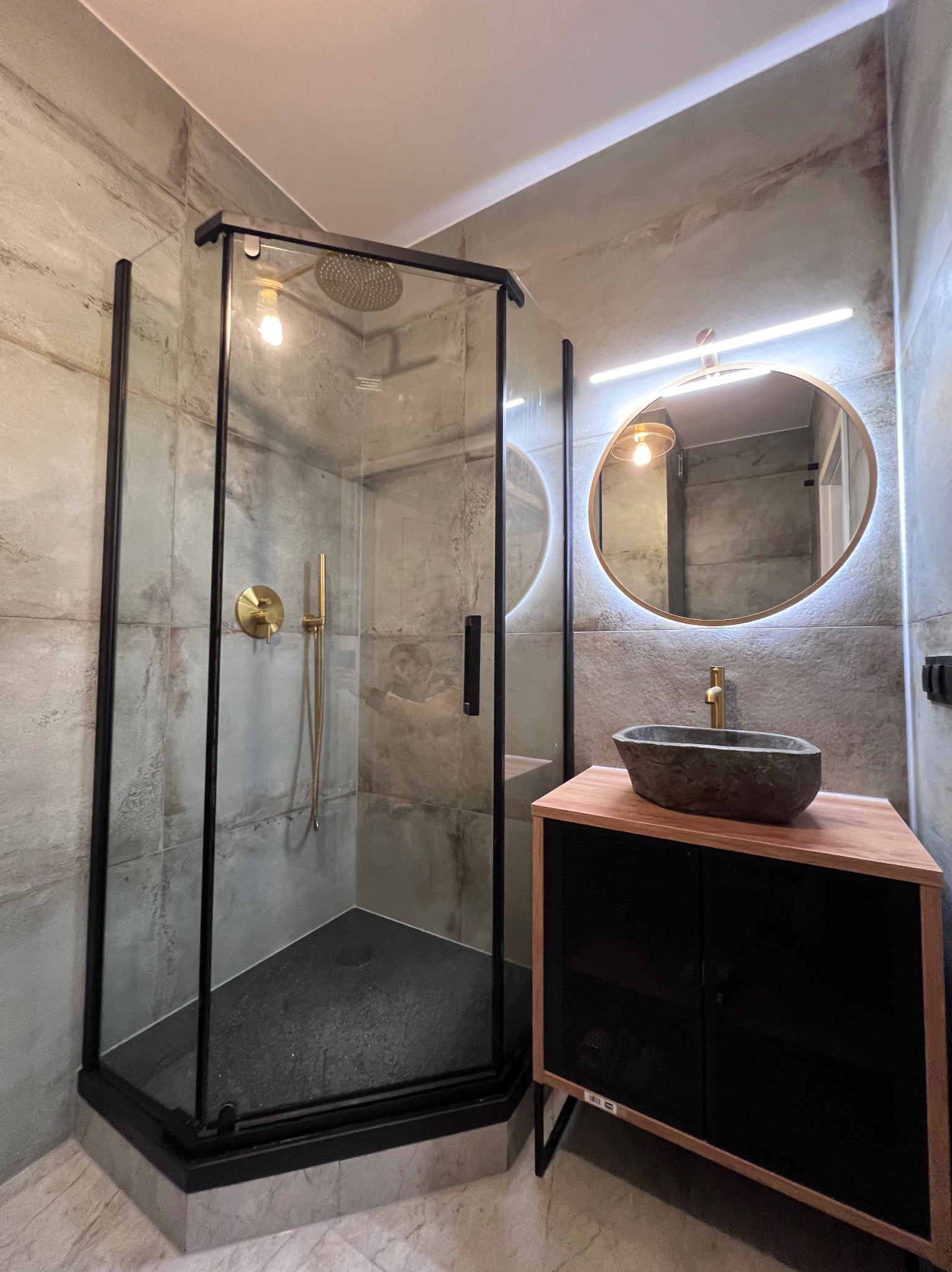 Nowoczesna łazienka z kabiną prysznicową narożną z czarnymi profilami, deszczownicą i baterią w kolorze złotym, umywalką w kształcie kamienia na drewnianej szafce oraz okrągłym lustrem...