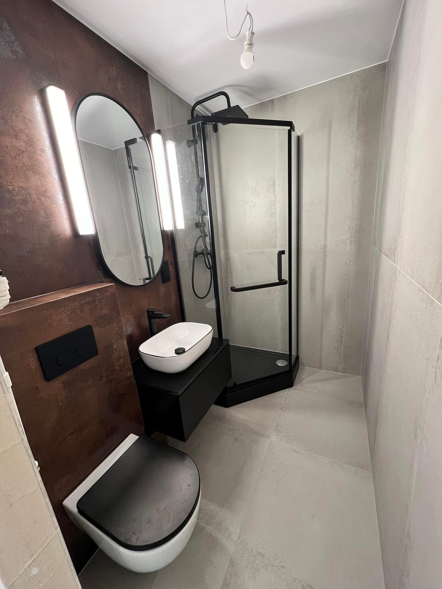 Nowoczesna łazienka z kabiną prysznicową z czarnymi ramami, owalnym lustrem podświetlonym ledami, umywalką nablatową i wiszącą toaletą. Ściany wykończone płytkami imitującymi beton i rdzę.