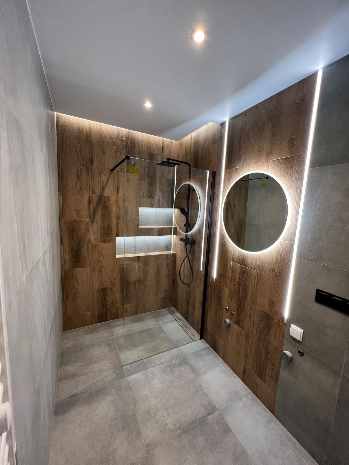 Nowoczesna łazienka z kabiną prysznicową typu walk-in, wykończona płytkami imitującymi drewno i szare płytki, z okrągłymi lustrami podświetlanymi LED i punktowym oświetleniem sufitowym.