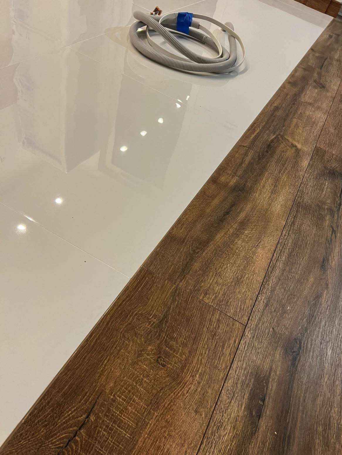 Połączenie błyszczących, białych płytek podłogowych i paneli podłogowych imitujących drewno, z widoczną rurą instalacyjną na płytkach.