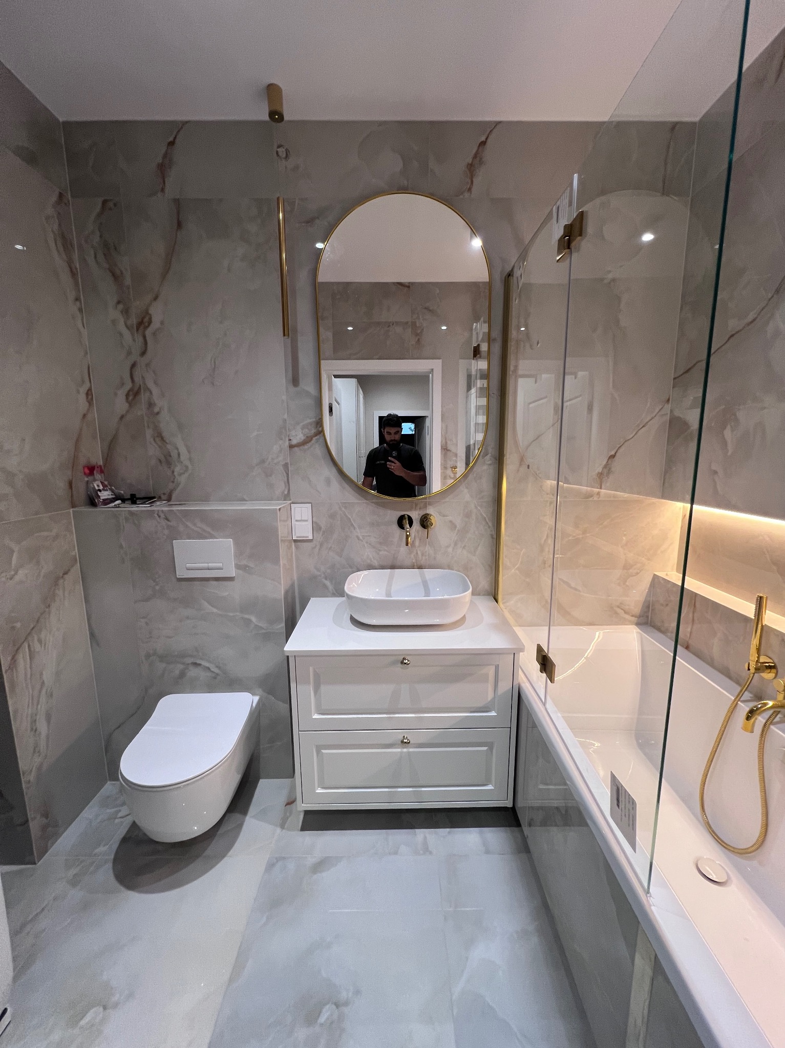 Nowoczesna łazienka z marmurowymi płytkami, owalnym lustrem w złotej ramie, białą umywalką nablatową na szafce z szufladami i kabiną prysznicową typu walk-in z złotą armaturą.