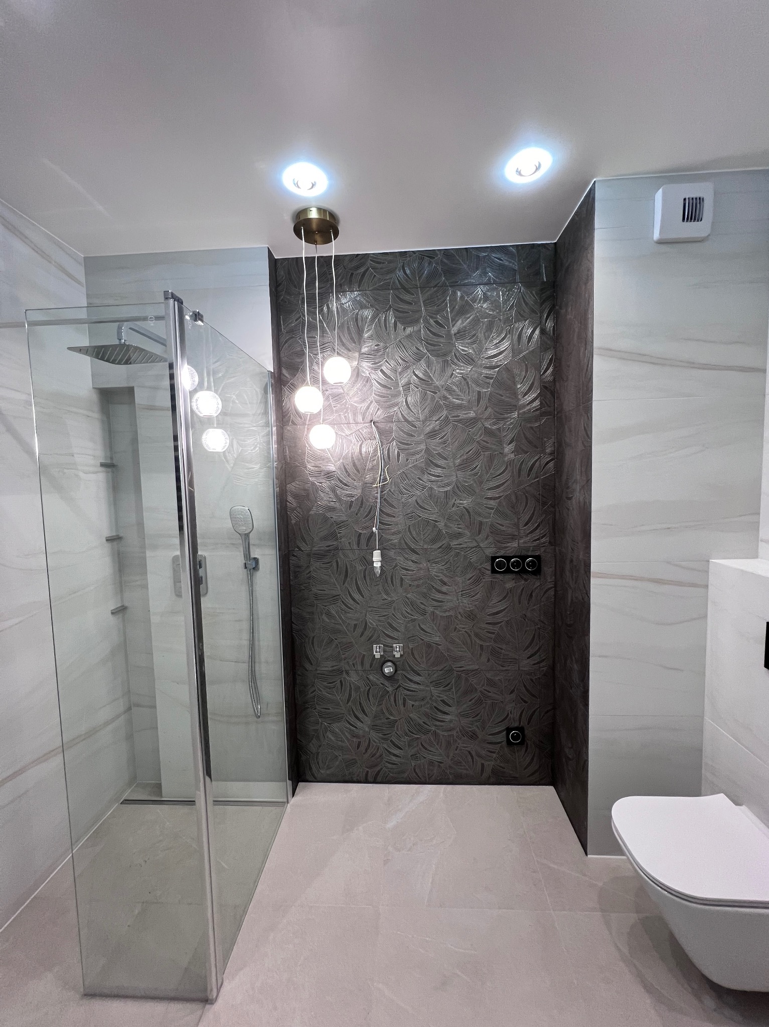 Nowoczesna łazienka z kabiną prysznicową typu walk-in, płytkami imitującymi marmur i dekoracyjną ścianą z motywem liści oraz wiszącą lampą z kulistymi kloszami.