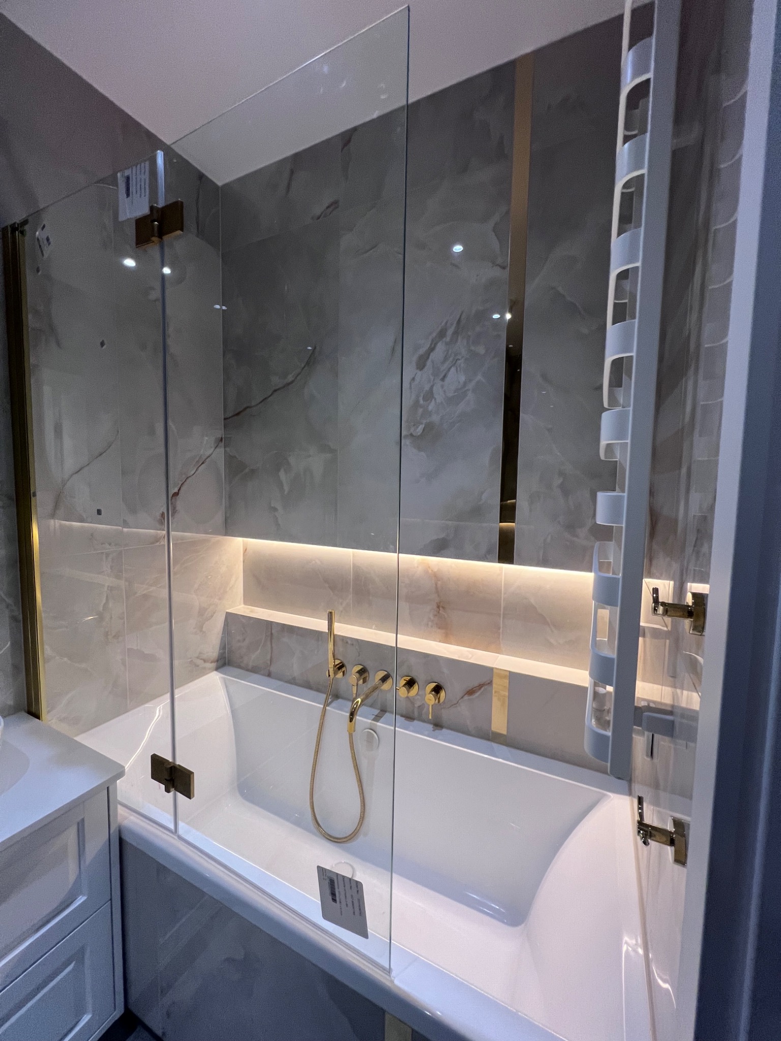 Nowoczesna łazienka z wanną obudowaną szklanym parawanem z mosiężnymi zawiasami, marmurowe płytki z podświetleniem LED, armatura w kolorze złotym, grzejnik drabinkowy.