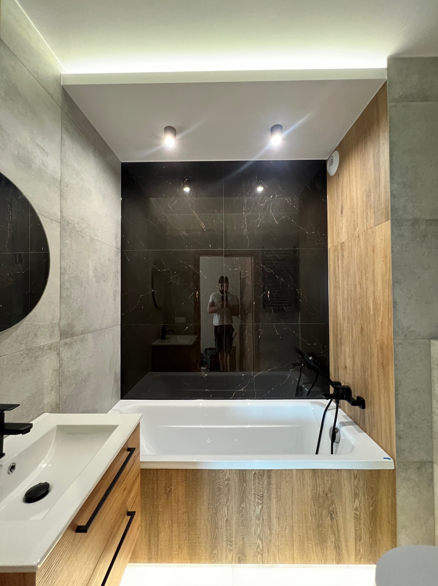Nowoczesna łazienka z czarnymi marmurowymi płytkami, drewnopodobnymi panelami, białą wanną i umywalką z drewnianą szafką, oświetleniem punktowym i ukrytym LED, widoczne odbicie osoby robiącej...