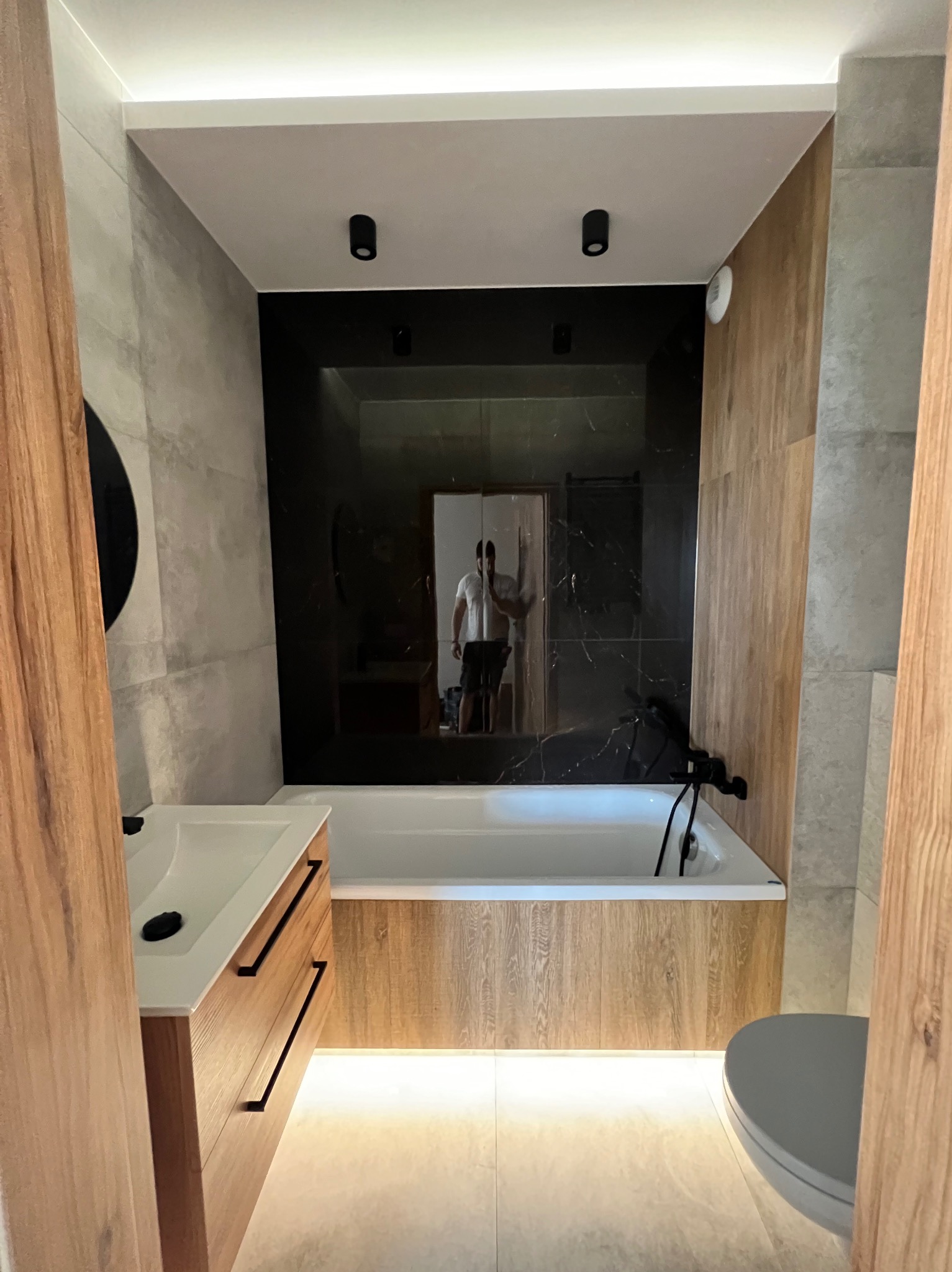 Nowoczesna łazienka z czarną armaturą, podświetleniem LED pod wanną i szafką umywalkową, oraz lustrzaną ścianą z odbiciem osoby.