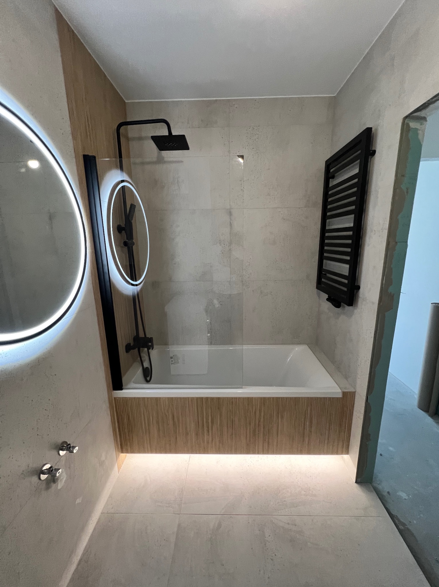 Nowoczesna łazienka z wanną za szklaną ścianką, czarną armaturą, podświetlanym lustrem i grzejnikiem drabinkowym, wykończona płytkami w odcieniach szarości i drewna.