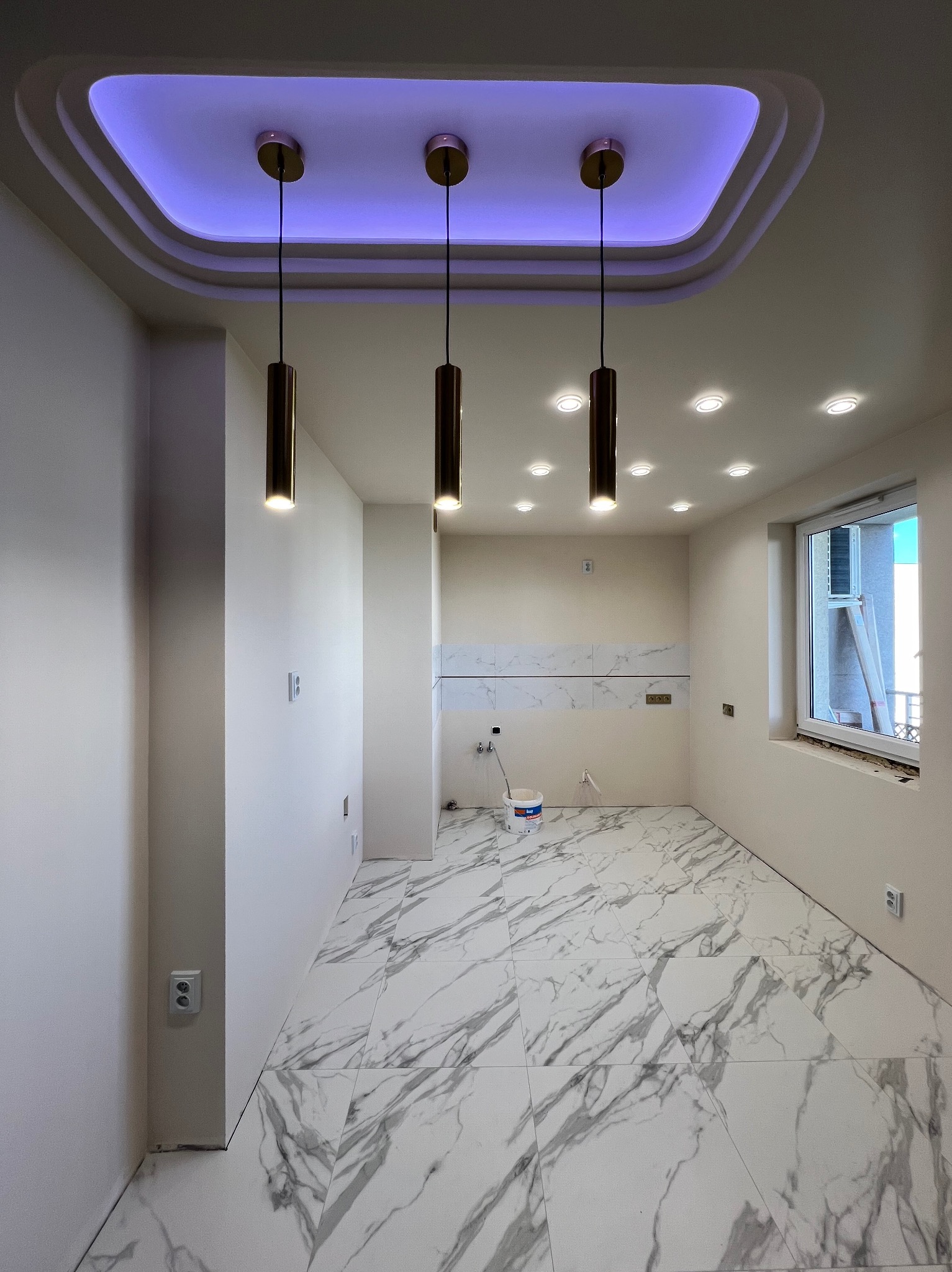 Wnętrze łazienki w trakcie prac wykończeniowych, widoczne marmurowe płytki na podłodze, fragment ściany z położoną glazurą, sufit z podświetleniem LED i trzema wiszącymi lampami, puste gniazdka...