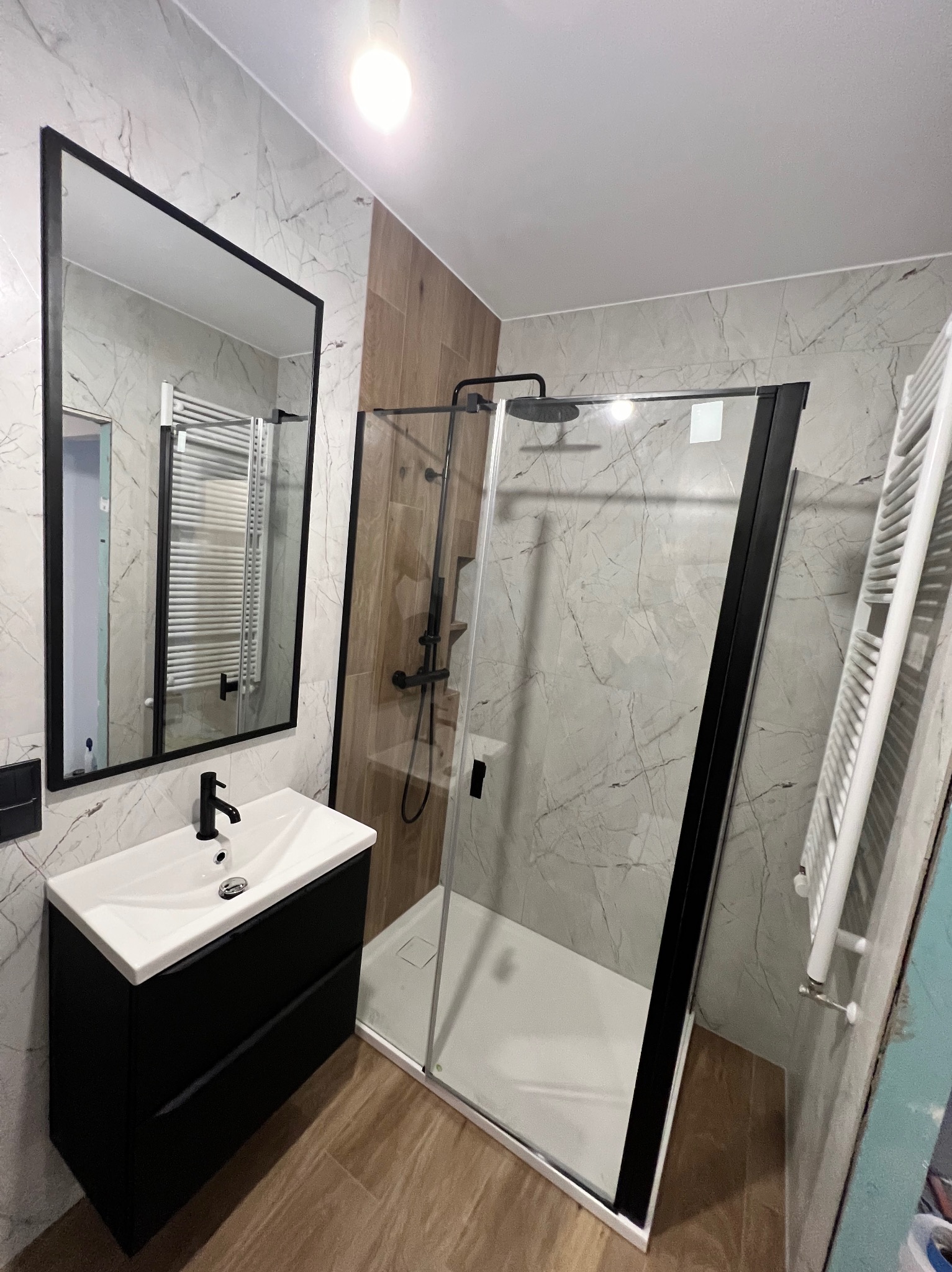 Nowoczesna łazienka z kabiną prysznicową typu walk-in z czarnymi profilami, czarną armaturą i umywalką z szafką w kolorze czarnym, wykończona płytkami imitującymi marmur i drewno.