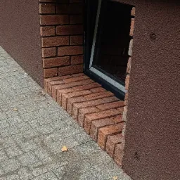 Okienko piwniczne z czarną ramą, obłożone czerwoną cegłą klinkierową, z brązowym tynkiem strukturalnym na ścianie i szarą kostką brukową na chodniku.