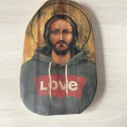 Ikona religijna przedstawiająca Jezusa w bluzie z kapturem i napisem 'LOVE' na czerwonym tle, umieszczona na drewnianej desce o nieregularnym kształcie.