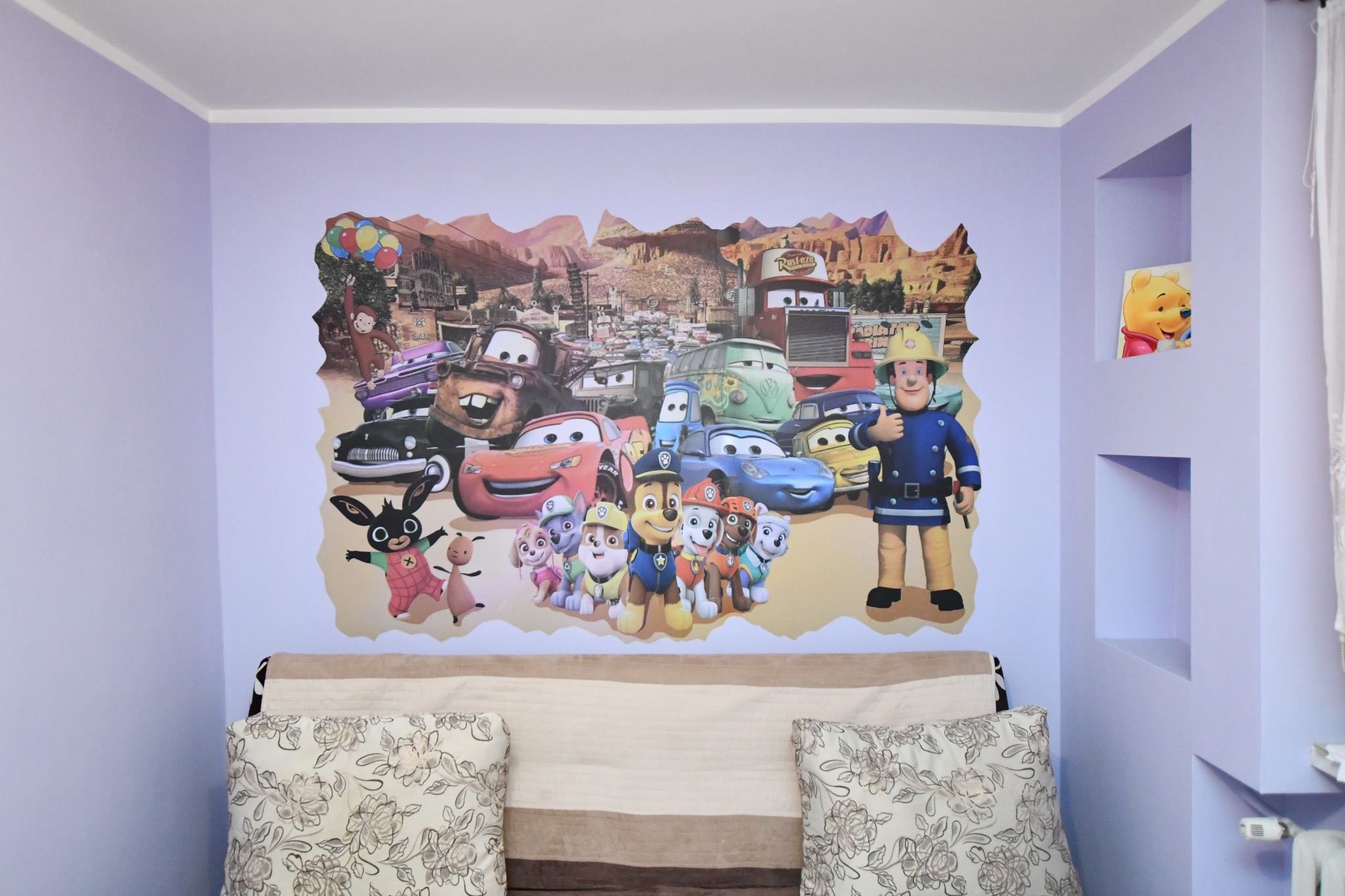 Dziecięcy pokój z fioletowymi ścianami, na których naklejona jest duża fototapeta z postaciami z bajek, nad łóżkiem z poduszkami w kwiaty, w ścianie wnęki z dekoracją.