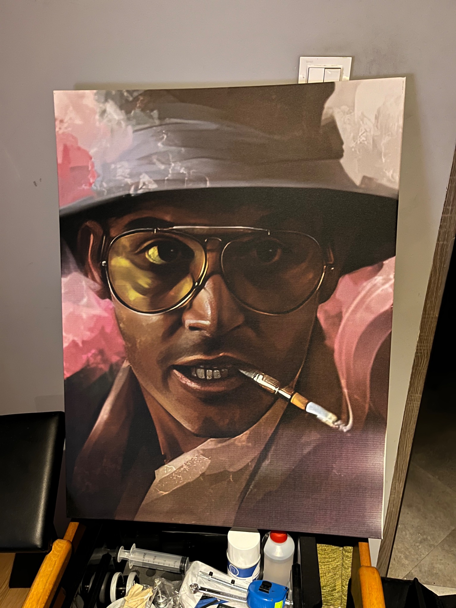 Portret w stylu pop-art przedstawiający mężczyznę w okularach i kapeluszu z papierosem w ustach, umieszczony na stojaku z widocznymi narzędziami malarskimi i akcesoriami.