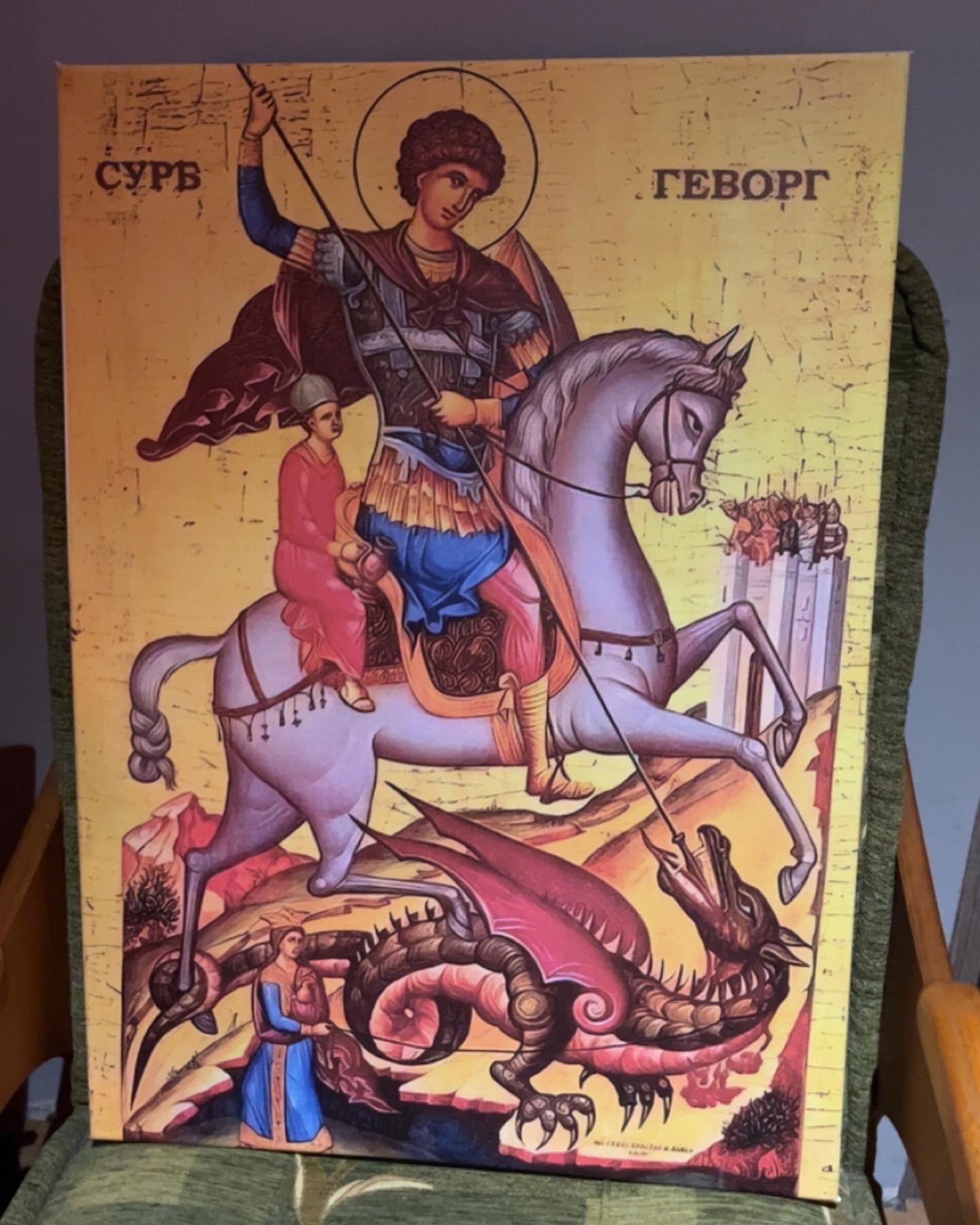 Ikona religijna przedstawiająca Świętego Jerzego zabijającego smoka, na płótnie z widoczną fakturą, ustawiona na zielonym fotelu.