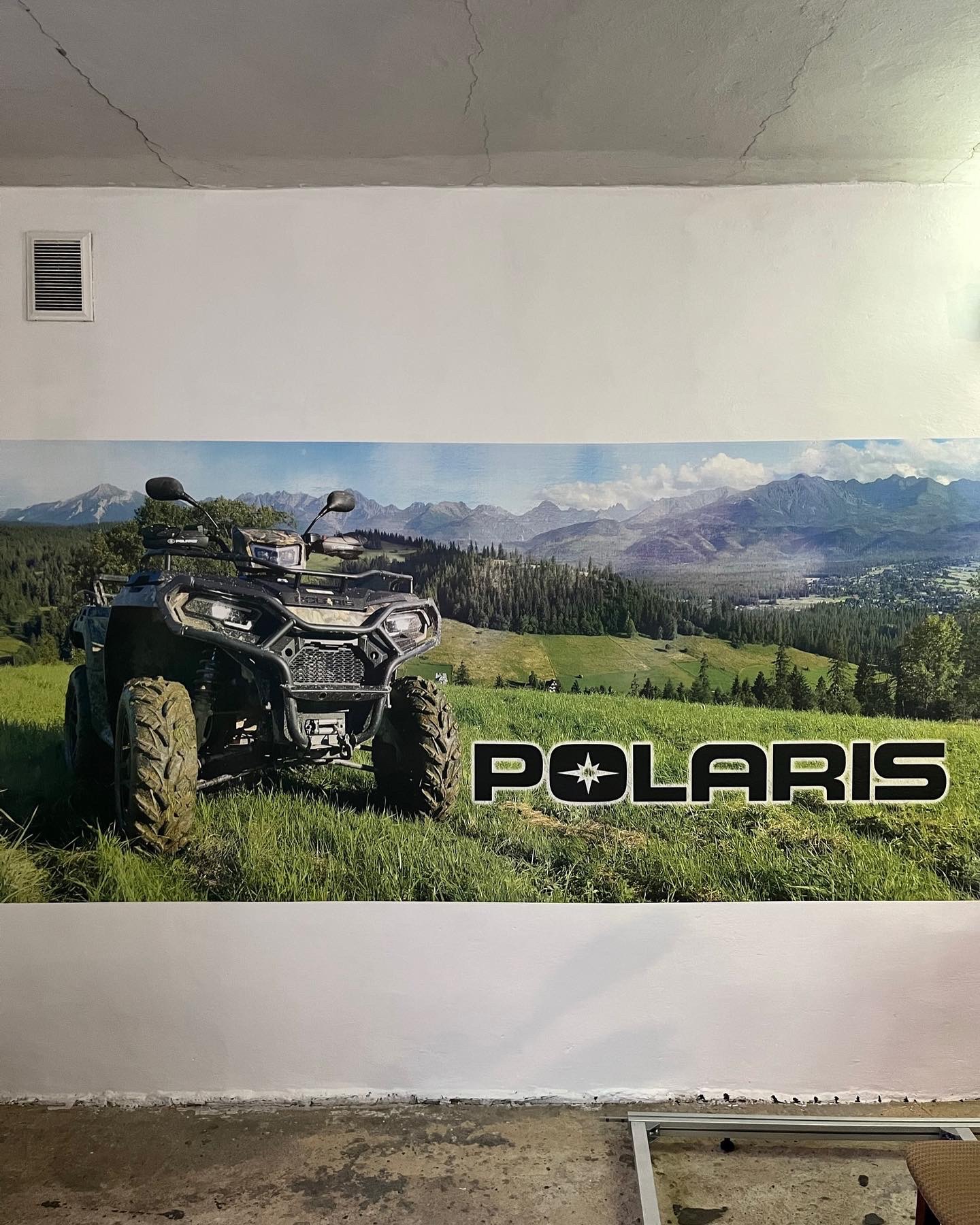 Duży plakat z pojazdem ATV marki Polaris na tle górskiego krajobrazu, naklejony na białej ścianie w pomieszczeniu z surowym wykończeniem.