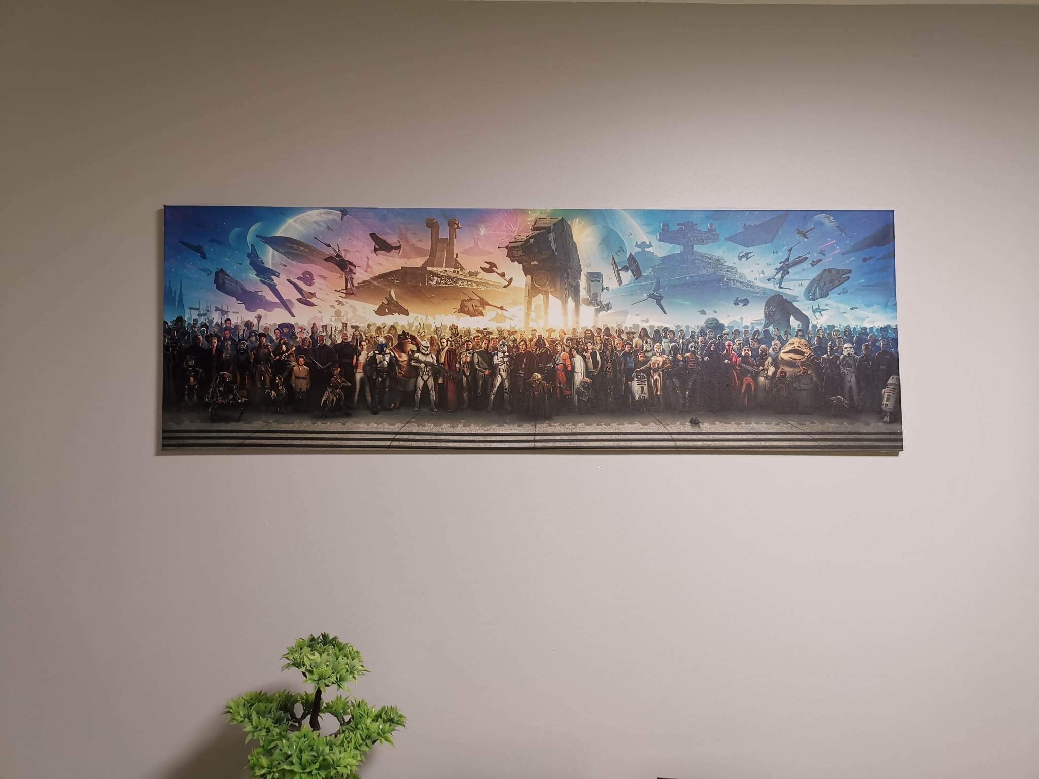 Panoramiczny plakat na ścianie przedstawiający postacie i statki z Gwiezdnych Wojen, umieszczony nad małym sztucznym drzewkiem bonsai.