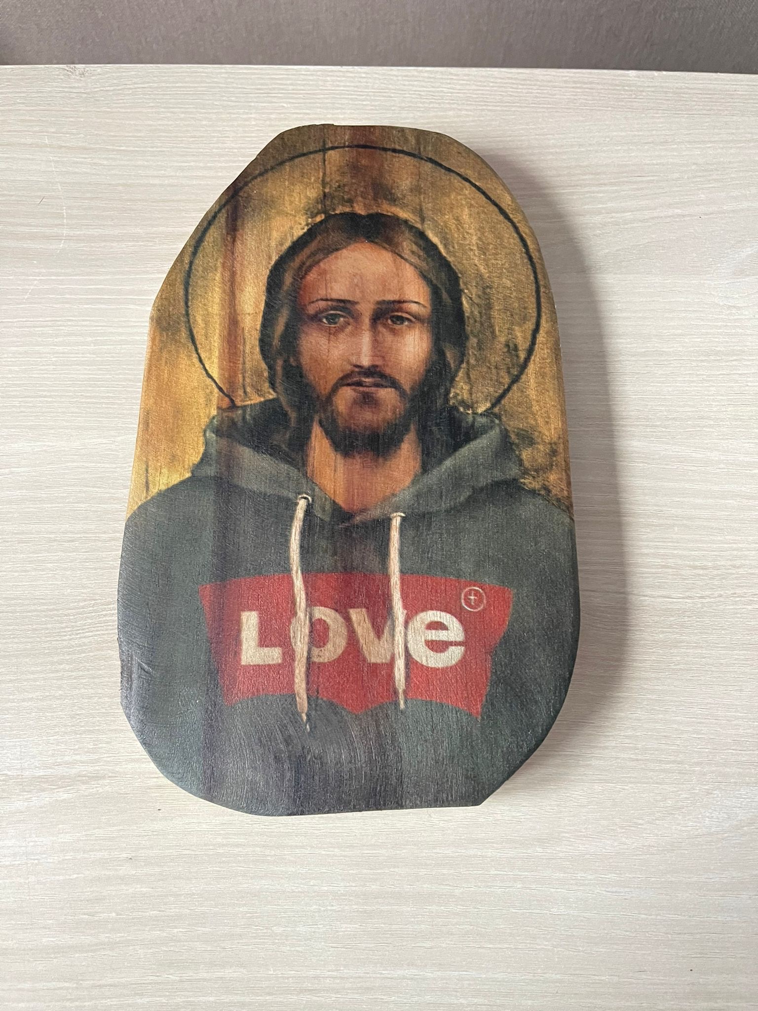 Ikona religijna przedstawiająca Jezusa w bluzie z kapturem i napisem 'LOVE' na czerwonym tle, umieszczona na drewnianej desce o nieregularnym kształcie.