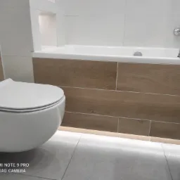 Nowoczesna, minimalistyczna łazienka z podwieszaną toaletą, obudową wanny imitującą drewno i szarą podłogą z delikatnym oświetleniem LED pod wanną.