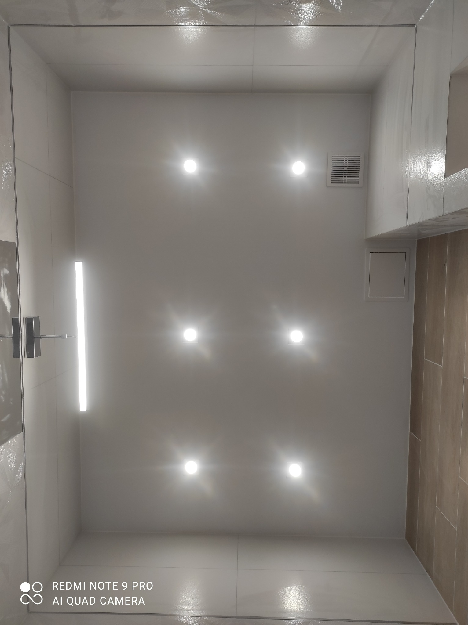 Wnętrze łazienki z widokiem na sufit z sześcioma punktowymi lampami LED i podłużną lampą liniową, białe płytki ścienne i podłogowe, kratka wentylacyjna, oraz brązowe płytki na jednej ze ścian.